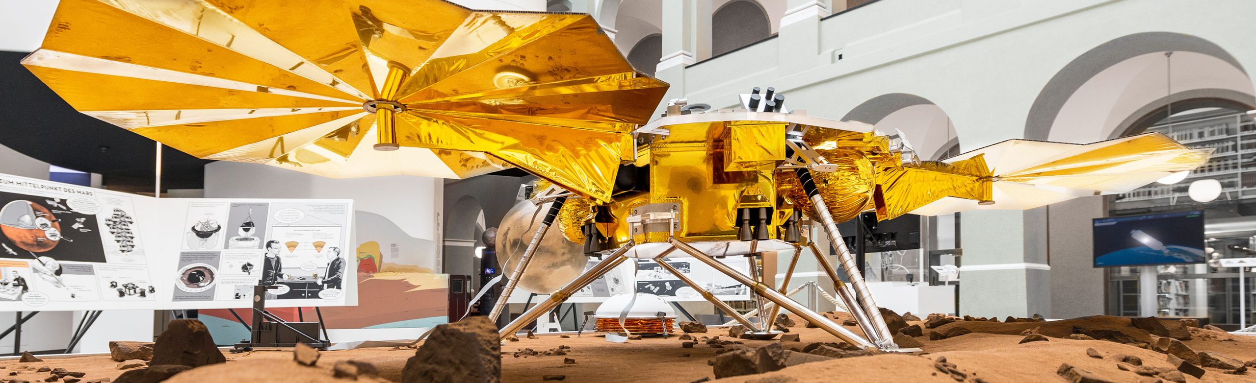 Exhibition model of a gold foil lander standing on simulated Martian soil inside a museum hall, shown on The Branco Weiss Fellowship website